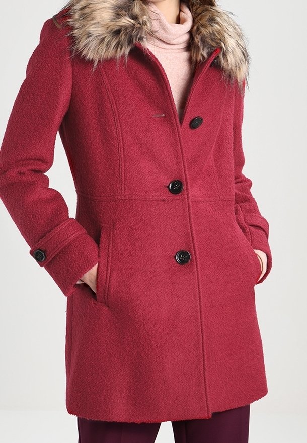 Manteau en laine rouge avec boutons noirs, poches latérales et col en fausse fourrure porté sur un col roulé rose clair.