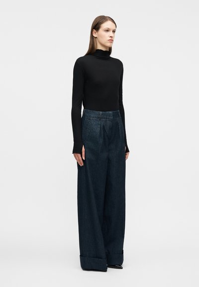 Γυναίκα που στέκεται, φοράει μαύρο μακρύ μανίκι ζιβάγκο και φαρδιά μπλε σκούρα τζιν με διπλωμένα τελειώματα, σε απλό λευκό φόντο.