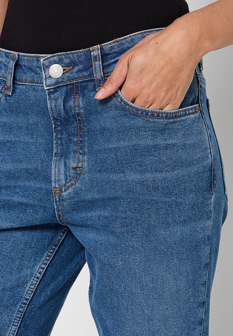 Hand rustend in de voorzak van blauwe denim jeans gedragen met een zwart top.