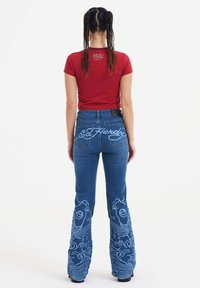 Rød tætsiddende skjorte med logo på ryggen; blå flagrende jeans med detaljeret fisk- og bølgebroderi. Sort mærke på livet.