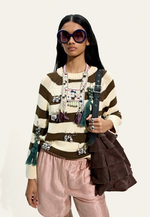Kobieta nosząca wielkie, okrągłe fioletowe okulary przeciwsłoneczne, pasiasty brązowo-kremowy sweter z frędzlami i dzwoneczkami, różowe szorty oraz niosąca dużą brązową zamszową torbę.