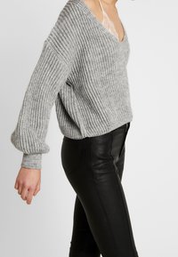 Grå stickad tröja med V-ringning, oversized passform och ribbad textur, tillsammans med svarta läderbyxor. Synliga manschetter och detaljer vid sömmen.