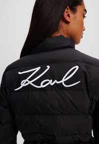 Schwarze gepolsterte Jacke mit hohem Kragen und weiß gesticktem "Karl"-Logo auf dem Rücken, mit einer glatten, glänzenden Textur.