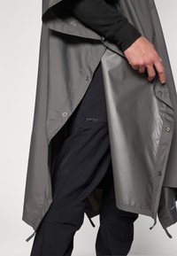Imperméable gris léger avec une texture lisse. Doté de fermetures à pression et d'un côté ouvert révélant un pantalon noir avec des poches zippées.