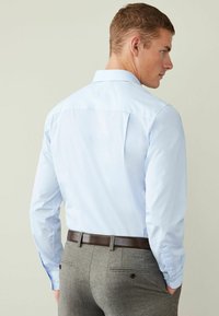 Helles blaues Langarmhemd mit Kragen, das ein Rückenpasse-Detail aufweist. Getragen mit grauen Hosen und einem braunen Leder-Gürtel.