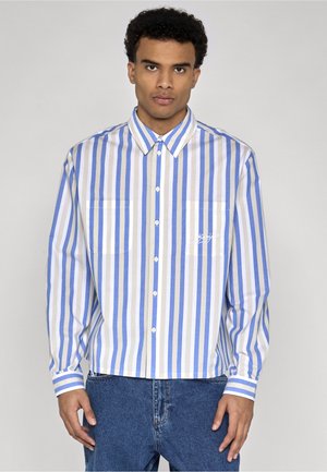 Jeune homme portant une chemise à boutons à rayures verticales bleues, blanches et beiges à manches longues, et un jean bleu, debout devant un fond uni.