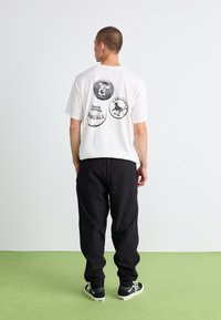 T-shirt alb din bumbac, cu trei imprimeuri grafice pe spate. Pantaloni negri, lejeri și adidași negri cu accente albe. Fundal simplu.
