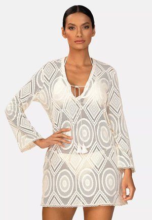 Kobieta w białej koronkowej sukience zakrywającej ciało z wzorami diamentów i kół, z frędzlami przy dekolcie i rękawami do połowy przedramienia.