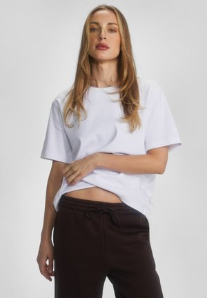 Kobieta o długich blond włosach, ubrana w biały t-shirt i ciemnobrązowe spodnie dresowe, stojąca na jasnym, jednolitym tle.