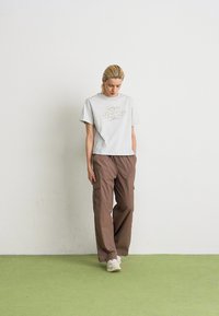 Femme aux cheveux blonds portant un t-shirt gris New Balance, un pantalon cargo brun ample et des baskets blanches, se tenant sur un sol vert contre un mur blanc.