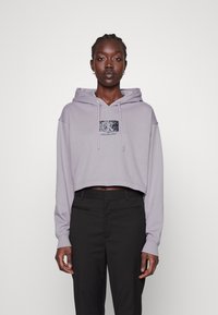 Jasnofioletowy krótki hoodie z kapturem ze sznurkiem i błyszczącą czarną łatą z logo. Połączony z czarnymi spodniami o dopasowanym kroju. Gładka tekstura materiału.