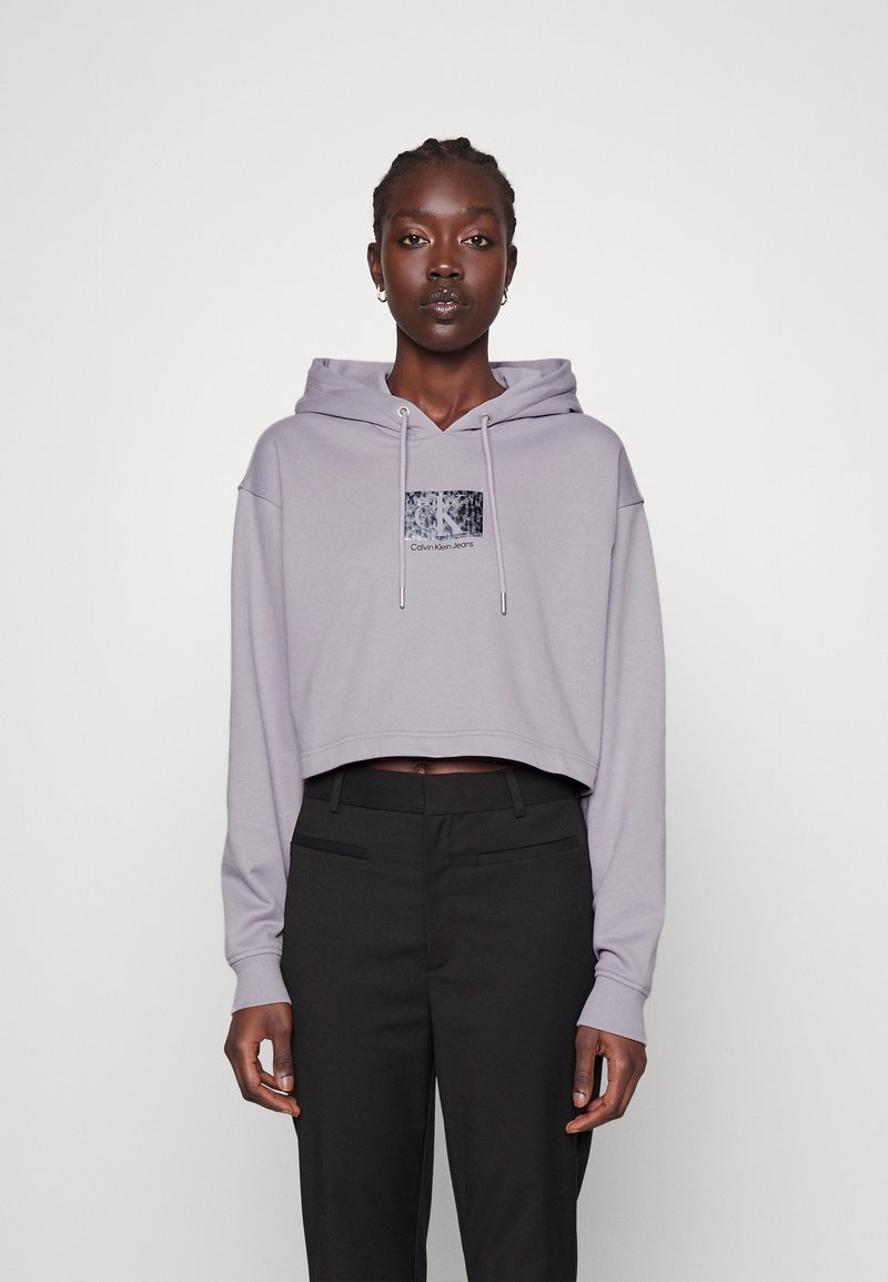 Jasnofioletowy krótki hoodie z kapturem ze sznurkiem i błyszczącą czarną łatą z logo. Połączony z czarnymi spodniami o dopasowanym kroju. Gładka tekstura materiału.