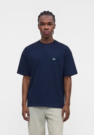 Ung mand med fletninger iført en mørkeblå Adidas T-shirt og lysegrå bukser, stående mod en ensfarvet lys baggrund.