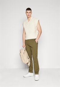Maglione senza maniche color crema con motivo a treccia, pantaloni oliva e zaino beige; sneakers bianche completano l'outfit.