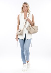 Witte mouwloze blazer, lichtblauwe skinny jeans en witte sneakers. Houdt twee beige handtassen vast, één met een schouderriem en één met handvatten.