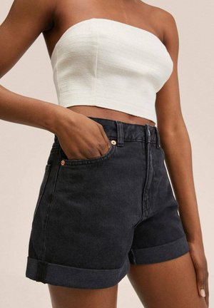 Person iført en hvid strapless crop top og sorte denimshorts med opkrullede kanter med den ene hånd i lommen mod en ensfarvet baggrund.