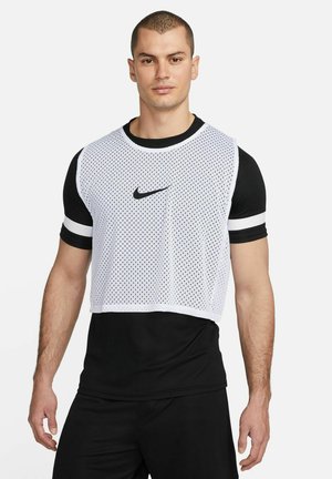 Un uomo sta in piedi con sicurezza, indossando una maglietta sportiva nera e una pettorina bianca in mesh con il logo Nike, su uno sfondo semplice.
