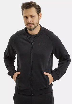 Mężczyzna z brodą w czarnej kurtce zapinanej na zamek, z rękami w kieszeniach, zwrócony twarzą do przodu na jednolitym białym tle.