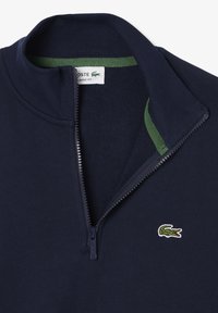 Navy pullover met een kwartrits, voorzien van een groene binnenkraag en een geborduurd krokodillologo op de borst. Glad, gestructureerd stof.
