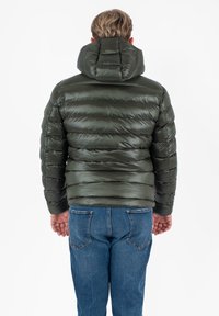 Man die met zijn rug naar de camera staat, gekleed in een donkergroene gewatteerde jas met capuchon en blauwe jeans, tegen een effen witte achtergrond.