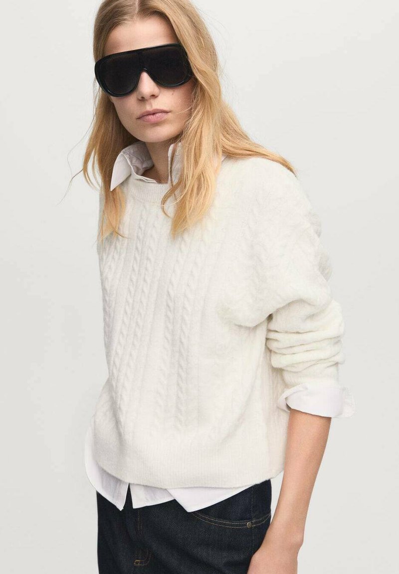 Maglione bianco a trecce con vestibilità rilassata, indossato sopra una camicia bianca con colletto. La modella indossa occhiali da sole neri oversize e jeans scuri.