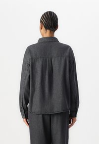 Camisa de denim preta, com um corte descontraído e mangas longas, punhos abotoados e detalhe de pregas nas costas. Textura suave e acabamento ligeiramente desgastado.