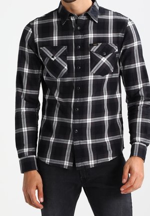 Uomo che indossa una camicia a quadri bianca e nera con due tasche sul petto, abbinata a un paio di jeans scuri.