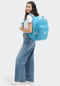 Ljusblå ryggsäck med flera fack, justerbara remmar och en nyckelringsaccessoar. Bärs av en person i avslappnad klädsel.