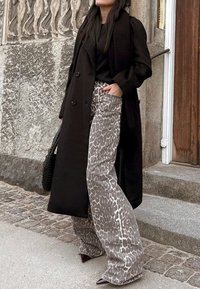 Garšs melns mētelis, pieguļošs melns tops un pelēkas leoparda raksta plaša kāju bikses. Stilīgas norādītās papēži un teksturēta melna soma. Akmens mūra fons.