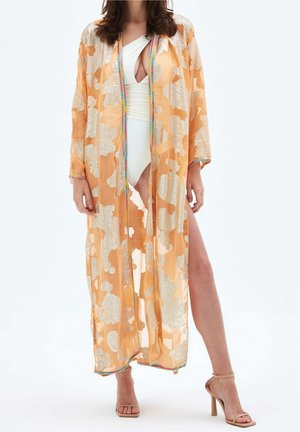 Femme portant un kimono floral orange transparent sur un maillot de bain une-pièce blanc avec une fente haute, associé à des sandales à talons hauts couleur nude.