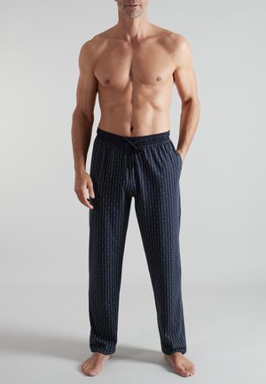 Oberkörperfreier Mann steht barfuß und trägt marineblaue Pyjamahosen mit kleinen weißen Punkten und einem elastischen Bund mit Kordelzug.