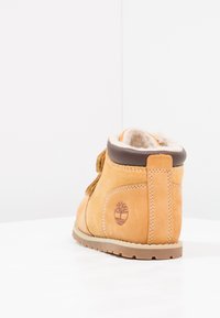 Stivaletto in suede beige con colletto imbottito marrone, dettagli cuciti e logo laterale. Presenta una suola in gomma e una fodera morbida.