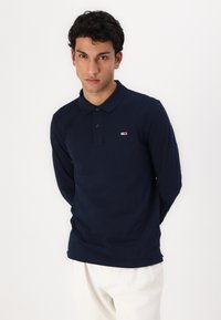 Polo a maniche lunghe blu navy con colletto e patta dei bottoni. Presenta un piccolo logo con accenti rossi e bianchi sul petto sinistro.