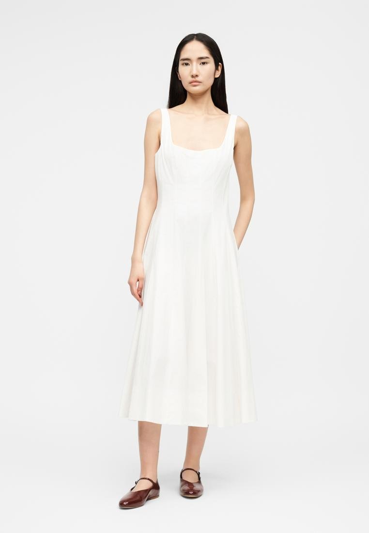 Femme aux longs cheveux noirs portant une robe midi blanche sans manches et des chaussures Mary Jane marron foncé, debout devant un fond gris uni.