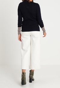 Marinblå ribbad tröja med flerfärgade randiga muddar, i kombination med vita vida byxor och olivgröna stövlar med rund detalj.