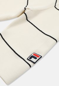 Kremowy dzianinowy sweter z cienkimi czarnymi poziomymi paskami i małą kwadratową naszywką z logo w kolorach czerwonym, białym i granatowym.