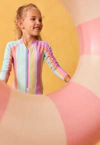 Maillot de bain à manches longues à rayures pastel dans des tons de rose, bleu et jaune avec une fermeture éclair devant. L'enfant se tient près d'une bouée rose et blanche.
