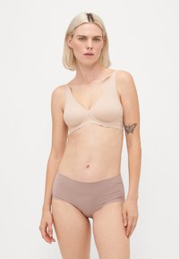 Woman with short blonde hair wearing a beige scalloped-edge bra and mauve seamless high-waist underwear, standing against a white background.