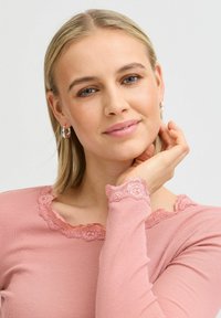 Rózsaszín, bordázott, hosszú ujjú felső csipkés díszítéssel a nyakán és az ujjak végén. Modell, hosszú szőke hajjal, aki a felsőt mutatja be, miközben az arcára támasztja a kezét.