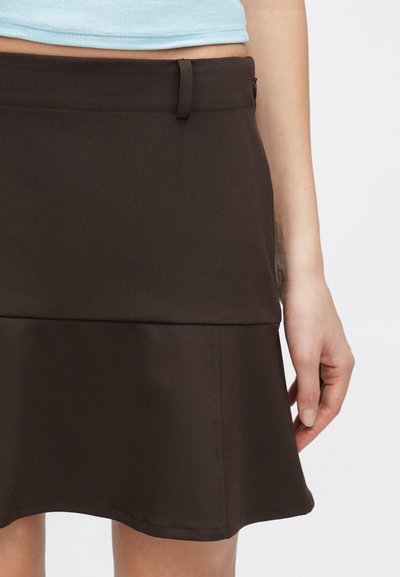 Jupe évasée marron à texture lisse, dotée d'une taille cintrée, d'une fermeture éclair latérale et d'un ourlet ondulé. Accentuée par une boucle de ceinture.