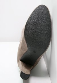 Botte en suède beige avec semelle noire texturée et petit talon, vue par le dessous sur fond blanc.