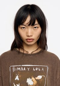 Pruun puuvillane ülekaaluline t-särk, millel on lillemotiividega trükise disain ja tekst "BIMBA Y LOLA." Kanda koos kuldse madu kaelakeega.