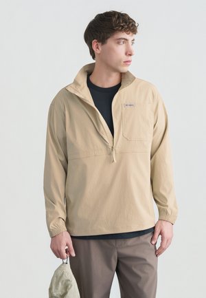 Jonge man draagt een beige quarter-zip jas over een zwart shirt en bruine broek, houdt een opgevouwen beige pet vast, kijkt naar rechts tegen een effen achtergrond.