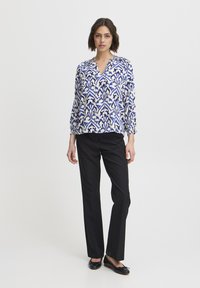 Bluza z vzorcem v modri in beli barvi z V-izrezom in nabranimi rokavi, kombinirana s črnimi oblikovanimi hlačami in črnimi ravnimi čevlji.
