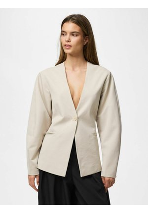 Vrouw draagt een beige colbert met één knoop en diepe V-hals en zwarte wijde broekspijpen, kijkt iets naar links.