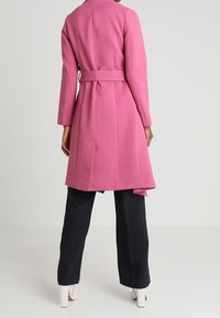 Langer pinker Mantel aus strukturiertem Stoff mit einem Gürtel in der Taille, ausgestattet mit einem taillierten Kragen und einem fließenden Rückenteil, kombiniert mit dunklen Hosen.