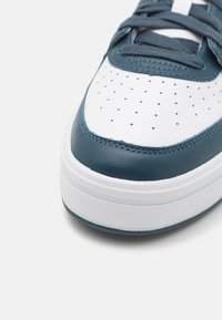 Zbliżenie na biały i ciemnoniebieski sneaker z perforowanymi detalami i teksturowaną gumową podeszwą na białym tle.