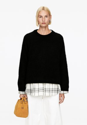 Schwarzer, flauschiger Strickpullover mit Rundhalsausschnitt, über einem weißen Karohemd getragen. Das Modell hält eine kleine, sandfarbene Wildledertasche. Weiße Hosen komplettieren den Look.
