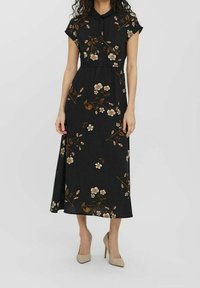 Svart midiklänning med korta ärmar, krage och beige blommönster, tillsammans med beiga högklackade skor mot en vit bakgrund.