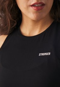 Haut noir sans manches en tissu lisse, arborant le mot « STRONGER » en blanc sur la poitrine. Design simple et ajusté.